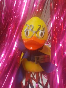 Rubber duck wearing glasses that say 2024, on a pink sparkly backdrop.