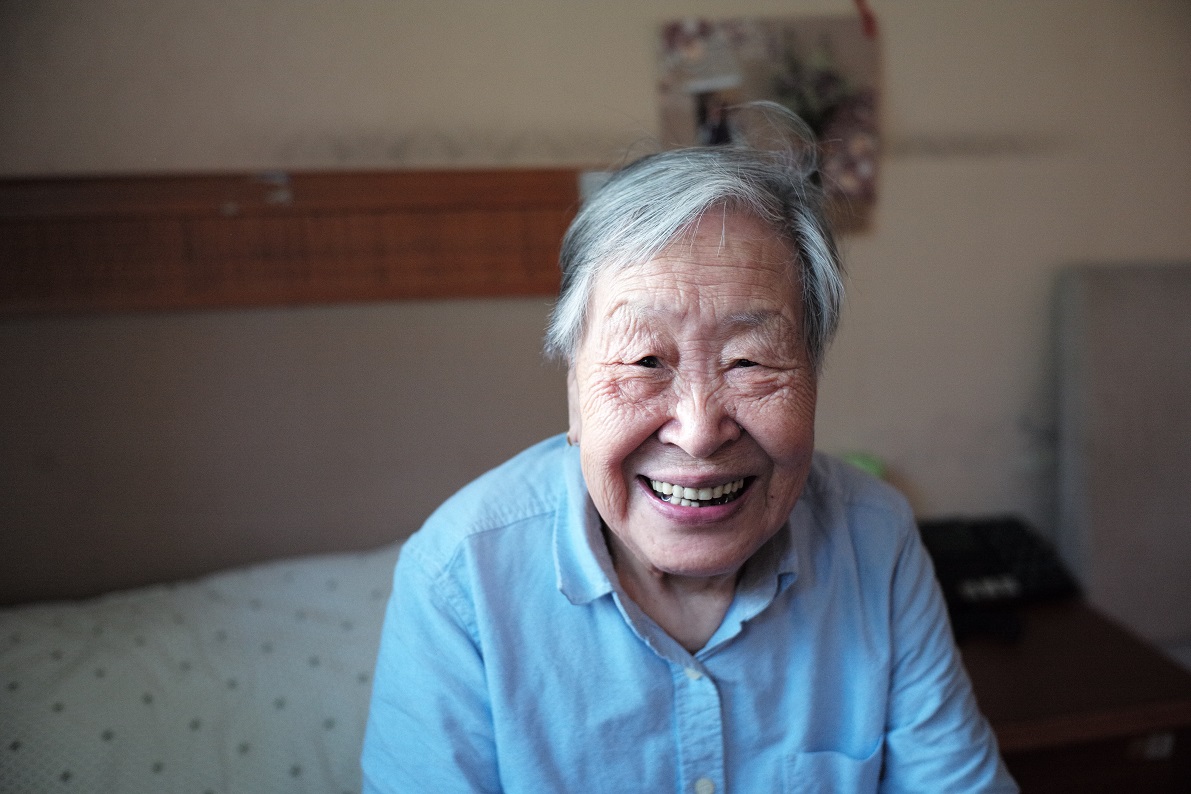 An older Asian man sits on his bed, smiling at the camera