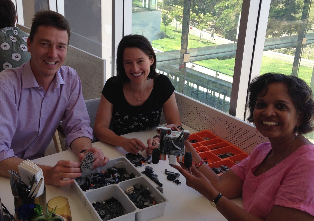 Dr David Hobbs, Associate Professor Belinda Lange, and Associate Professor Pammi Raghavendra seated around a table, looking at the LEGO Mindstorm E3 robot.