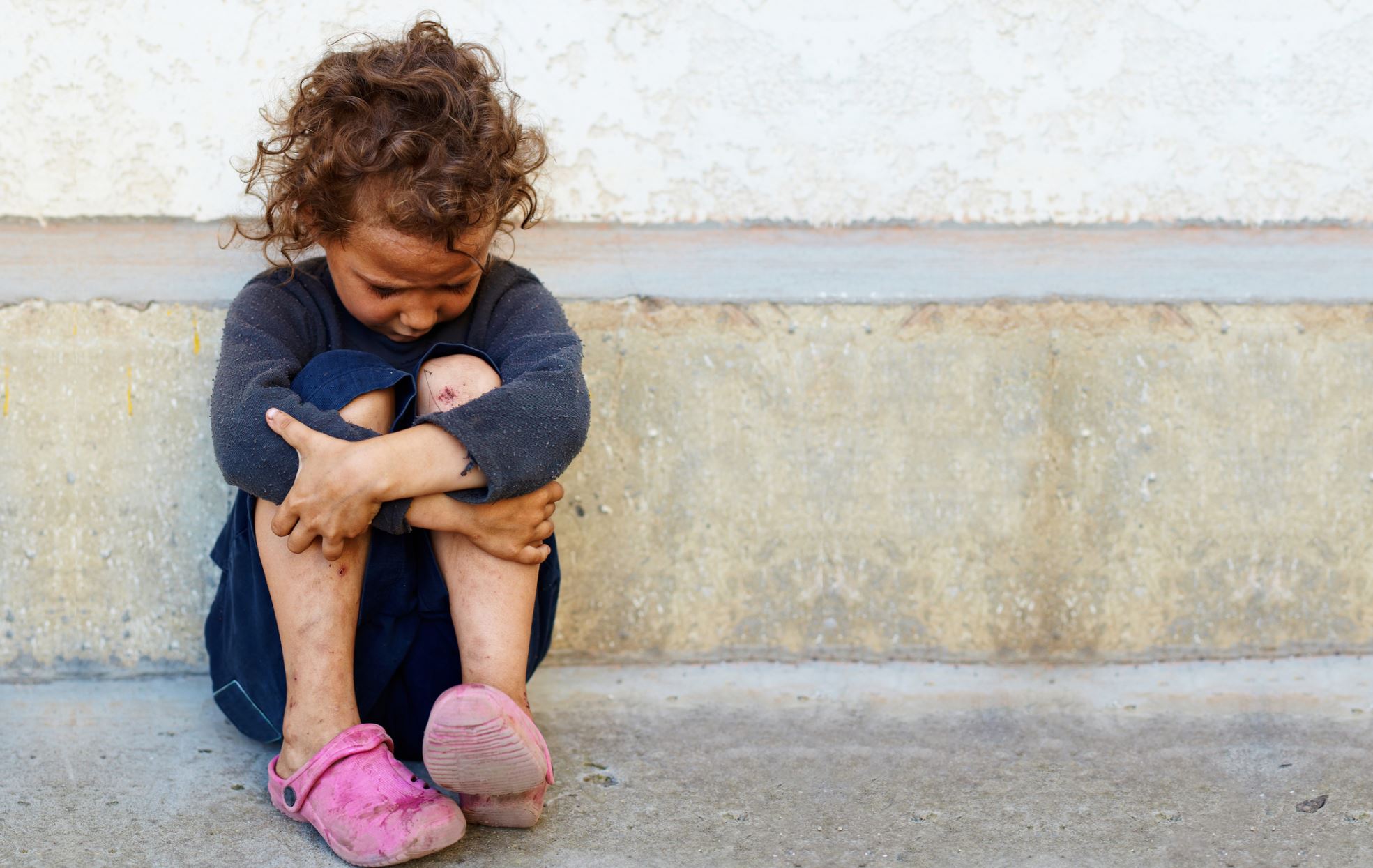A dirty child in old clothing sits dejectedly on a concrete step. She looks scared and sad.