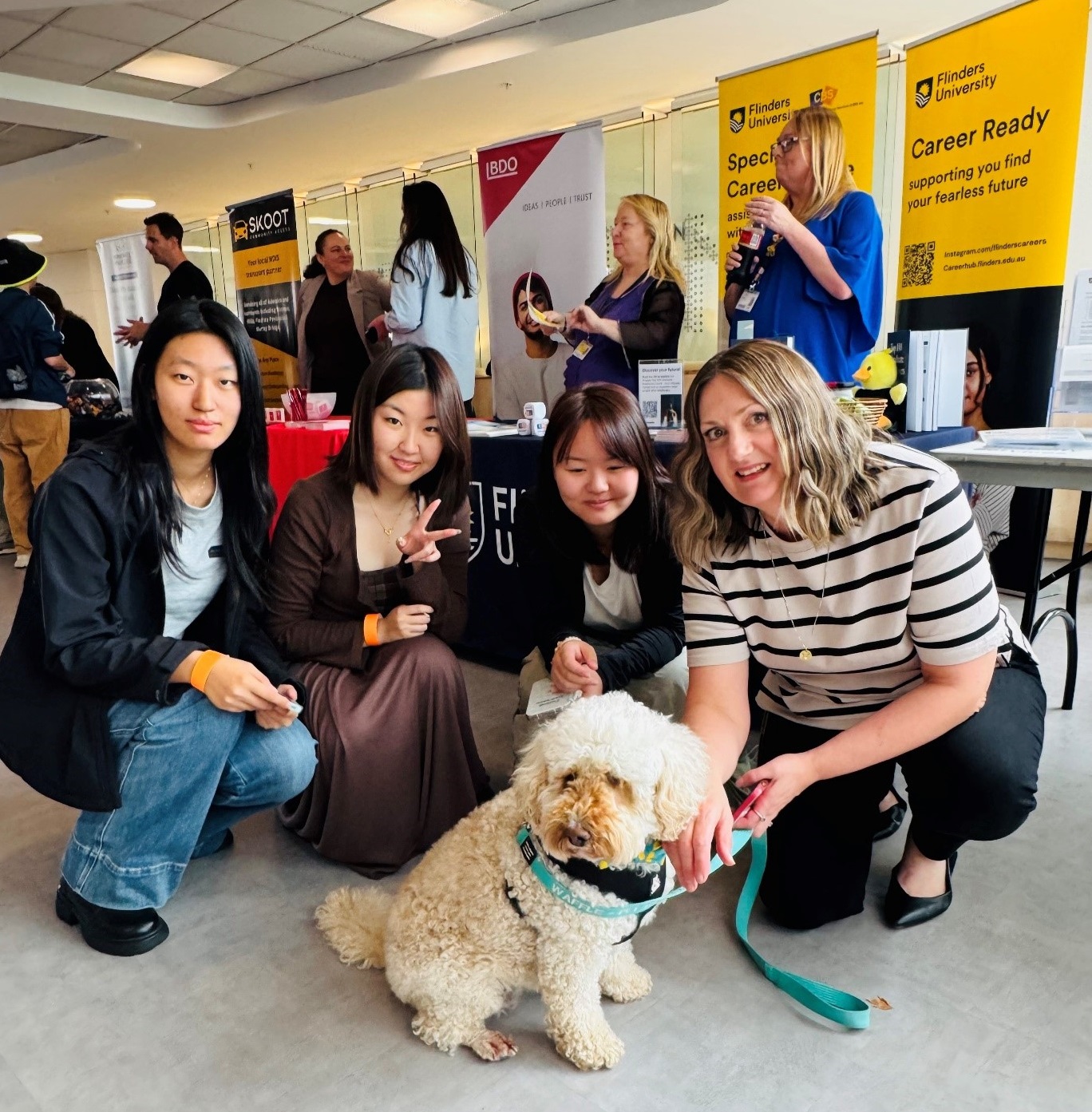 Flinders Inclusion Event photo. Four women squatting down with a Cavoodle support dog. Exhbitors standing at the trestles behind them.