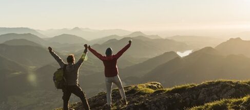 Two hikers on top of a mountain with their hands raised in triumph.
